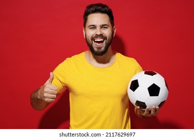 Smiling fun young bearded man football fan in yellow t-shirt cheer up support favorite team hold in hand soccer ball showing thumb up like gesture isolated on plain dark red background studio portrait