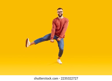 Smiling funky cheerful young bearded man with short hair in glasses wearing red sweatshirt and denim pants dancing on one leg isolated on yellow background. Good mood happy lifestyle nice day concept.