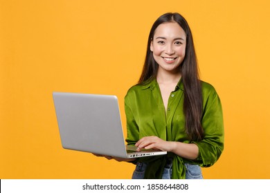 Smiling funny beautiful young brunette asian woman wearing casual basic green shirt standing working on laptop pc computer looking camera isolated on bright yellow colour background, studio portrait