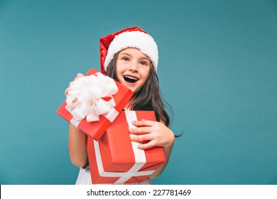 Smiling Funny Child (kid, Girl) In Santa Red Hat. Holding Christmas Gift In Hand. Christmas Concept. Shooting On Blue Background