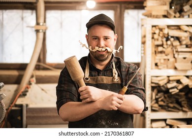 Smiling, funny male carpenter in cap and protective coveralls with shaving chip mustache, holding chisel, mallet crossed