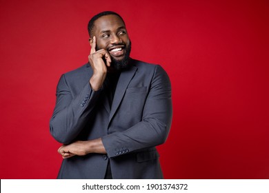 Smiling Funny Pensive Young African American Business Man 20s Wearing Classic Jacket Suit Standing Put Hand Prop Up On Chin Looking Aside Isolated On Bright Red Color Wall Background Studio Portrait