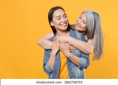 Smiling Funny Pretty Family Asian Women Gray-haired Mother And Brunette Daughter In Casual Clothes Posing Hugging Looking At Each Other Isolated On Yellow Color Wall Background Studio Portrait