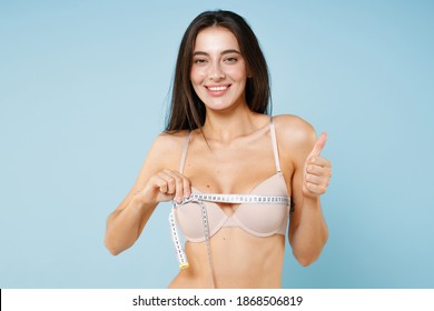 Smiling funny young brunette woman 20s in beige brassiere underwear standing posing measuring breast with measure tape showing thumb up isolated on pastel blue colour wall background studio portrait