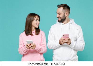 Smiling funny young couple two friends man woman 20s wearing white pink casual hoodie using mobile cell phone typing sms message isolated on blue turquoise colour wall background studio portrait