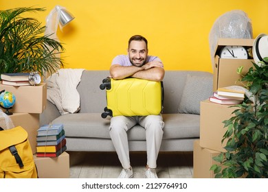 Smiling funny young owner man hold suitcase sits in living room on sofa at home household unpacking stuff indoor plant rents flat isolated on yellow wall. Relocation moving in new apartment concept