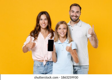 Smiling Funny Young Parents Mom Dad With Child Kid Daughter Teen Girl In Basic T-shirts Hold Mobile Phone With Blank Empty Screen Showing Thumbs Up Isolated On Yellow Background. Family Day Concept