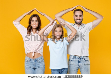 Smiling funny young parents mom dad with child kid daughter teen girl in basic t-shirts holding hands above head like roof of house isolated on yellow background studio portrait. Family day concept