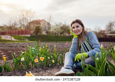 Smiling gardener relaxing among fresh tulips, daffodils, hyacinths in spring garden. Happy woman admires colorful flowers sitting on the ground - Powered by Shutterstock