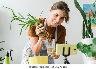 Smiling gardener wearing gloves holding plant roots and recording video blog about repotting plants using smartphone at home - Powered by Shutterstock