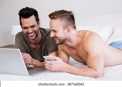 Smiling Gay Couple Using Laptop And Phone On Bed