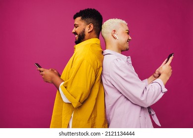 Smiling Gay Couple Using Mobile Phones In A Studio. Happy Young Couple Texting While Standing Back To Back Against A Purple Background. Cute Gay Couple Enjoying Being Online.