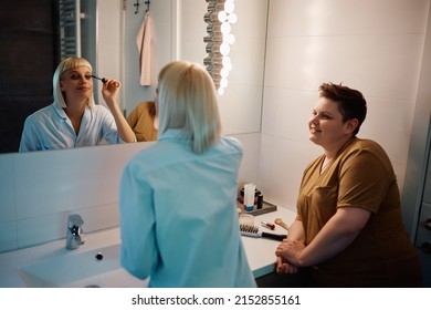 Smiling Gay Woman Looking At Her Girlfriend Who Is Applying Make-up In Front Of Bathroom Mirror. 