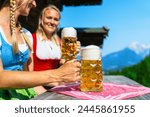 smiling german girls in traditional dirndls or tracht toasting beer mugs in front of the bavarian alpine mountains behind a alpine cabin. Oktoberfest concept image