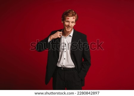 Smiling ginger man with red lipstick marks on face posing isolated over red studio wall