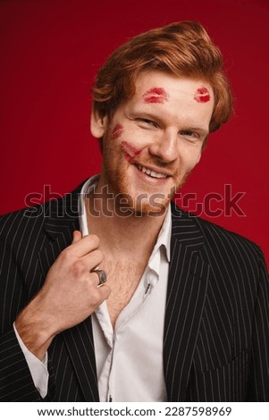 Smiling ginger man with red lipstick marks on face posing isolated over red studio wall