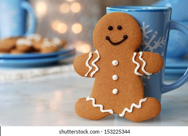 Smiling Gingerbread Man Standing Next To Snowflake Mug.  Plate Of Additional Cookies And Defocused Holiday Lights In Background.  Closeup With Shallow Dof.  Copy Space Included For Text.