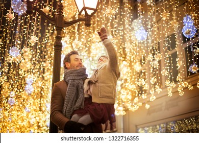 smiling girl in the arms of the father in a brown coat pulls his hands to the lights on the decorated street of the city center - Powered by Shutterstock