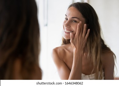 Smiling girl in bath towel look in mirror apply moisturizing facial cream or serum, happy young woman perform daily morning skincare face routine for healthy glowing skin, do beauty procedure