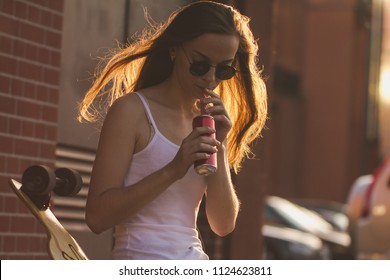 A smiling girl in black sunglasses with a longboard, has a rest on the street in the evening and drinks a cocktail. - Powered by Shutterstock