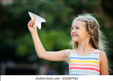 Smiling Girl In Bright Dress Playing With Flying Paper Airplane Toy Outdoors