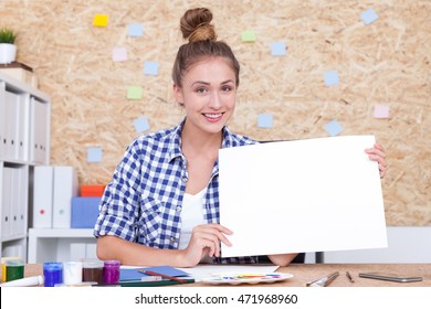 Smiling Girl With Bun Is Sitting In Office And Holding Large Piece Of Paper. Concept Of Advertisement. Mock Up