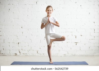 Smiling Girl Child Practicing Yoga, Standing In Vrksasana Exercise With Namaste, Tree Pose, Working Out Wearing Sportswear, T-shirt, Pants, Indoor Full Length, White Loft Studio Background 