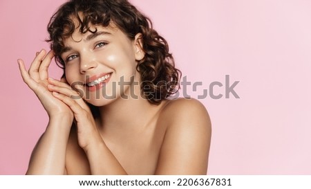 Smiling girl with curly hair, nourished clean body and face, touching her soft skin after shower, cosmetic treatment, pink background.