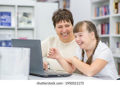 Smiling girl with down syndrome is uses a laptop with her teacher at library. Education for disabled children concept