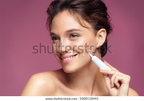 Smiling girl with flawless\
skin applying treatment cream on pink background. Skin care\
concept