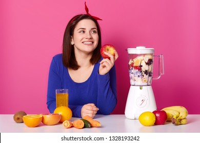 Smiling Girl With Fresh Fruits On Table Isolated Over Pink Background. Lady Looks Aside While Making Coktail. Woman Holds Apple In Left Hand Thinks About Something Pleasent. Health Concept.