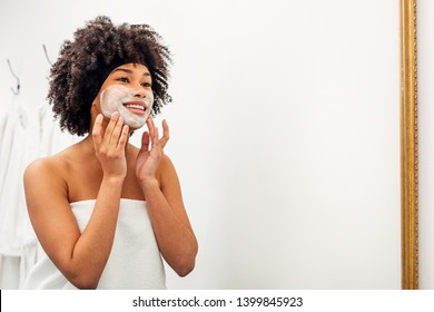 Smiling girl in front of a mirror. Young woman applying white natural mask with hands.