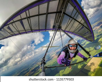 Smiling girl hang glider pilot shows thumb up while flying high over green fields below clouds. Wide angle selfie photo of extreme sport taken with action camera
