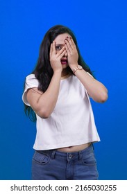 Smiling girl, hiding face behind palms, looking with one eye. Isolated over blue background. Beautiful amazed Indian girl or Young south Asian woman hiding eyes and face and looking with only one eye