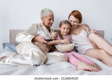 Smiling Girl Holding Popcorn While Watching Tv Near Lesbian Moms At Home