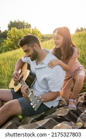 Smiling Girl Hugging Shoulders Of Dad Playing Guitar While Resting In Countryside