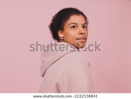 Smiling girl is listening music. Close up portrait. Young black woman in wireless headphones against pink background. Minimal style