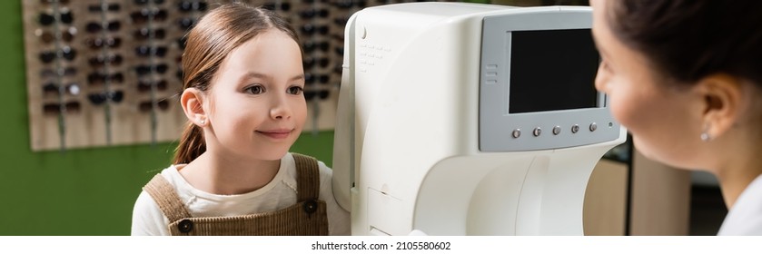 smiling girl looking at blurred oculist near vision screener in optics salon, banner