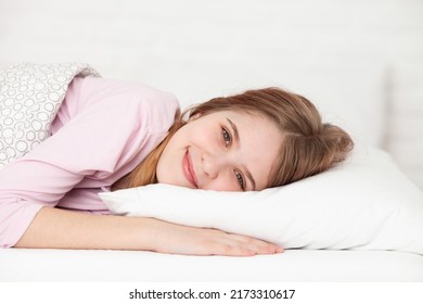 Smiling Girl Lying On A White Pillow In Bedroom. Cute Teen Girl Relaxing After Waking Up At Home. Healthy Sleep, Good Morning Concept. Copy Space. Selective Focus.