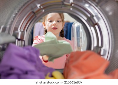 Smiling girl in pigtails unpacks washing machine, takes out colorful wet clean smelling clothes from the drum, spending time in the laundry room household chores.