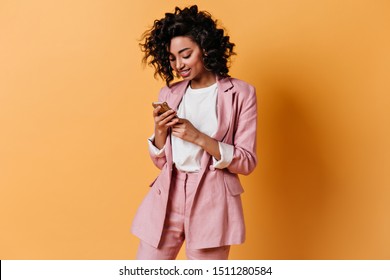 Smiling girl in pink jacket texting message. Glad young woman in suit holding smartphone.