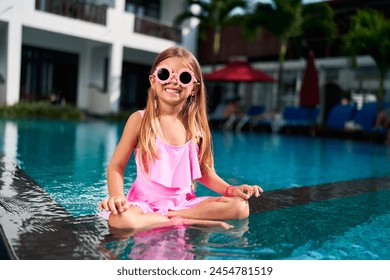 Smiling girl in pink swimsuit enjoys summer by pool. Happy child with sunglasses sits at water edge, legs dipped in. Kid relaxing, having fun at tropical resort on sunny day. Playful vacation moments. - Powered by Shutterstock