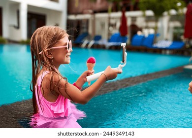 Smiling girl in pink swimsuit takes selfie in pool, holding ice cream. Child enjoys summer day, captures moment with smartphone on selfie stick. Bright, sunny leisure activity, kid at resort. - Powered by Shutterstock