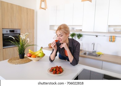 Smiling girl poses on camera in excellent spirits with bright ripe strawberry and tastes berries, standing in stylish and bright kitchen with white cabinets, household appliances and variety of