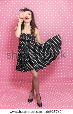 Smiling girl is posing in beautiful dotted dress and hiding one eye behind a tasty pink doughnut.