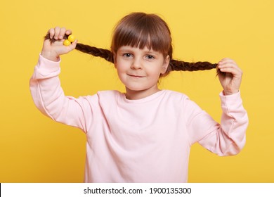 Smiling Girl Pull Her Pigtails Up By Hand And Looking Directly At Camera Isolated Over Yellow Background, Charming Female Kid Having Fun Alone, Child In Pale Pink Casual Shirt.