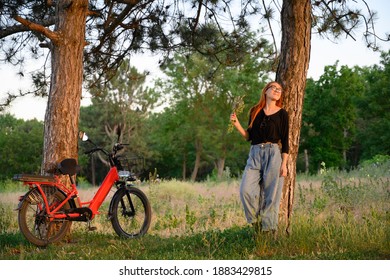 Smiling Girl With Red Electro Bicycle Standing Near Tree At Background Of Green With Bouquet Of Wild Flowers. Enjoy After Bike Ride At Sunset Summer Day. Recreation, People, Healthy Lifestyle Concept