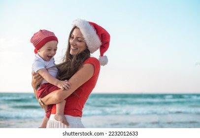 Smiling girl in red shirt and Santa hat holds her son on beach. woman and child laughing and joy Christmas. Celebrate holiday together. have fun on holiday and vacation - Powered by Shutterstock