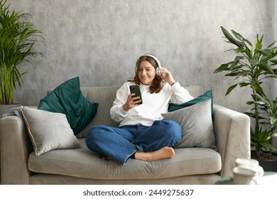 Smiling girl relaxing at home, she is listening to music, using a smartphone and wearing white headphones. Young woman hearing enjoying good audio sound lounge on couch at home - Powered by Shutterstock