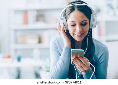 Smiling girl relaxing at home, she is playing music using a smartphone and wearing white headphones